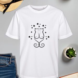 Bijela majica s crnim printom dviju stiliziranih mačaka koje sjede leđima jedna uz drugu, s repovima oblikovanim u elegantne spirale koje tvore srce. Oko njih su raspoređena mala crna srca, dodajući romantičan i minimalistički dojam. Majica je položena na ravnu površinu uz slameni šešir s crnom vrpcom, svijeću, zelenu biljku u tegli, otvorenu knjigu i traperice.