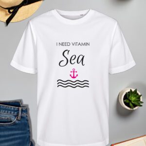 Bijela majica s natpisom "I NEED VITAMIN SEA" u crnoj boji, gdje je riječ "Sea" ispisana elegantnim rukopisnim fontom. Ispod natpisa nalazi se ilustracija ružičastog sidra i stiliziranih crnih valova, što stvara morski i opuštajući dojam. Majica je položena na ravnu površinu uz slameni šešir s crnom vrpcom, svijeću, zelenu biljku u tegli, otvorenu knjigu i traperice.
