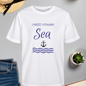 Bijela majica s natpisom "I NEED VITAMIN SEA" u plavoj boji, gdje je riječ "Sea" ispisana elegantnim rukopisnim fontom. Ispod natpisa nalazi se ilustracija crne sidra i stiliziranih valova u plavoj boji, što stvara morski i opuštajući dojam. Majica je položena na ravnu površinu uz slameni šešir s crnom vrpcom, svijeću, zelenu biljku u tegli, otvorenu knjigu i traperice.