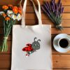 Platnena vrećica s ilustracijom simpatične bubamare u crveno-crnim krilima i tenisicama, koja leti i veselo maše. Vrećica je položena na drveni stol, okružena buketima tratinčica, narančastih cvjetova i lavande, uz šalicu crne kave na bijelom tanjuriću.