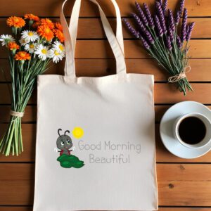 Platnena vrećica s natpisom *"Good Morning Beautiful"*, ilustracijom simpatične bubamare koja sjedi na listu i veselo maše, uz sunce koje sja. Vrećica je položena na drveni stol, okružena buketima tratinčica, narančastih cvjetova i lavande, uz šalicu crne kave na bijelom tanjuriću.