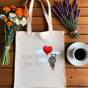 Platnena vrećica s natpisom "Love is in the Air" i ilustracijom simpatične bubamare koja drži crveni balon u obliku srca, okružena oblacima. Vrećica je položena na drveni stol, uz bukete tratinčica, narančastih cvjetova i lavande, te šalicu crne kave na bijelom tanjuriću.