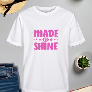 bijela majica s natpisom "MADE TO SHINE" u ružičastoj boji, ukrašenom zvjezdicama oko riječi "TO". Majica je položena na ravnu površinu uz slameni šešir s crnom vrpcom, svijeću, zelenu biljku u tegli, otvorenu knjigu i traperice.