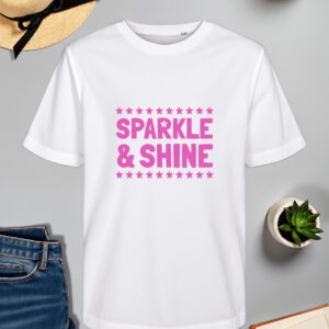 Bijela majica s natpisom "SPARKLE & SHINE" u ružičastoj boji, okruženom redovima malih zvjezdica. Majica je položena na ravnu površinu uz slameni šešir s crnom vrpcom, svijeću, zelenu biljku u tegli, otvorenu knjigu i traperice.