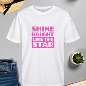 Bijela majica s natpisom "SHINE BRIGHT LIKE THE STAR" u ružičastoj boji, s dizajnom unutar okvira i zvjezdicom unutar slova "A". Majica je položena na ravnu površinu uz slameni šešir s crnom vrpcom, svijeću, zelenu biljku u tegli, otvorenu knjigu i traperice.