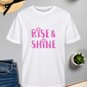 Bijela majica s natpisom "Rise & Shine" u ružičastoj boji, ukrašenim zvjezdicama. Majica je položena na ravnu površinu uz slameni šešir, svijeću, zelenu biljku u tegli, otvorenu knjigu i traperice.