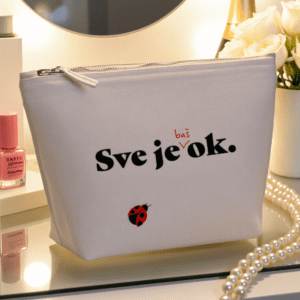 Kozmetička torbica u bež boji s natpisom **"Sve je baš ok."** i ilustracijom bubamare, smještena na elegantnom toaletnom stolu uz bisernu ogrlicu, lak za nokte i bijele ruže.