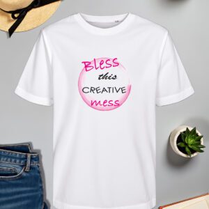 Bijela majica s natpisom *"Bless this creative mess"*, gdje su riječi ispisane u kombinaciji crnog i ružičastog fonta. Dizajn je upotpunjen akvarelnim ružičastim krugom koji daje umjetnički i razigrani izgled. Majica je položena na ravnu površinu uz slameni šešir s crnom vrpcom, svijeću, zelenu biljku u tegli, otvorenu knjigu i traperice.