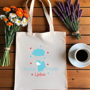 Bež platnena vrećica s tiskanim dizajnom leži na drvenoj podlozi. Dizajn prikazuje plavog anđela s aureolom, koji visi s plavog oblaka poput padobrana, okruženog malim crvenim srcima i plavim zvjezdicama. Riječi 'Radost' i 'Veselje' napisane su svijetloplavom bojom, dok je 'Ljubav' ispisana crvenom. Lijevo od vrećice nalazi se buket bijelih tratinčica i narančastog cvijeća,