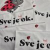 Bijele majice s tiskom natpisa "Sve je baš OK" i motivom bubamare raspoređene na drvenoj površini. Jedna majica sadrži ilustraciju simpatične bubamare s osmijehom i podignutom rukom. Dizajn kombinira crnu i crvenu boju, stvarajući upečatljiv vizualni identitet.