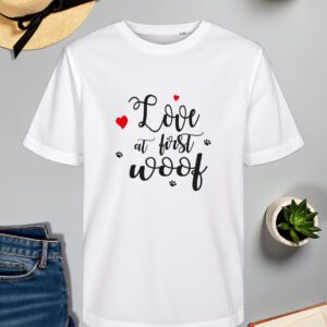 Bijela majica s natpisom "Love at first woof", ispisanim elegantnim crnim fontom, ukrašena crvenim srcima i otiscima psećih šapa. Majica je položena na sivoj površini, okružena modnim i dekorativnim detaljima – slamnatim šeširom s crnom vrpcom, trapericama, upaljenom svijećom, sukulentom u bijeloj posudi i otvorenom knjigom.